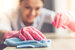 Woman in protective gloves is smiling and wiping dust using a spray and a duster while cleaning her house, close-up